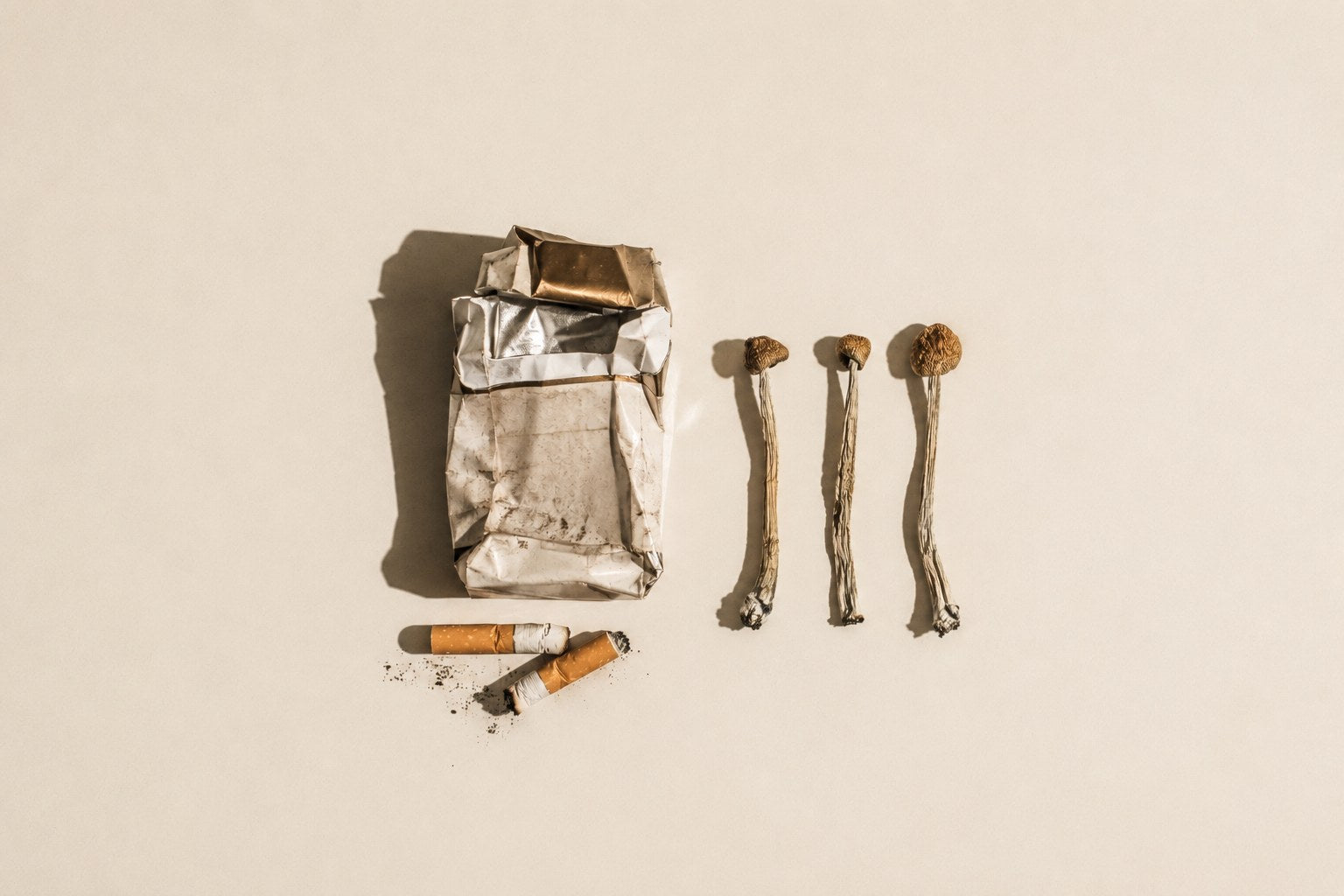 Overhead still life: a crushed cigarette packet and two stubbed-out cigarettes on the left, three dried psilocybe mushrooms in a row on the right