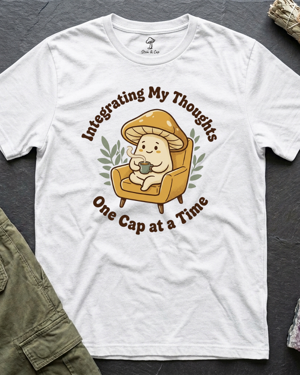 Integrating My Thoughts tee in white, a mushroom settled into an armchair with a hot drink, shot flat on slate
